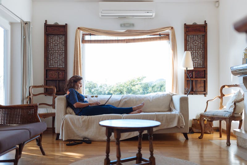 woman sits on sofa, in front of a large window, in living room with wooden decor