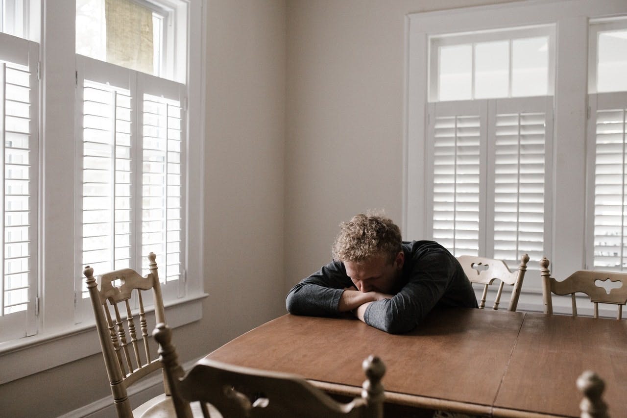 Man with stressed sad expression in grey jumper sits with head resting on wooden kitchen table 