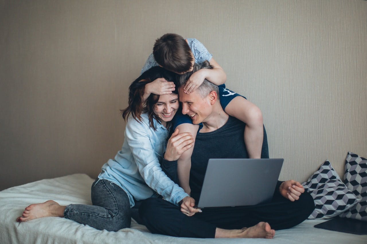 A family sat on a bed laugh and hug while looking at a laptop