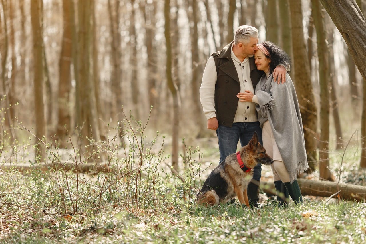 An older couple hug in a forest, a german shepherd sits next to them