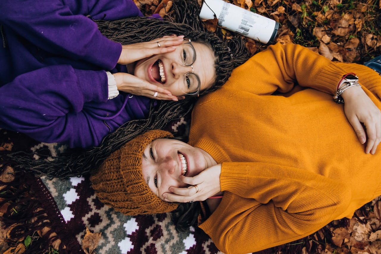 two friends with excited expressions and colourful jumpers lie on a rug 