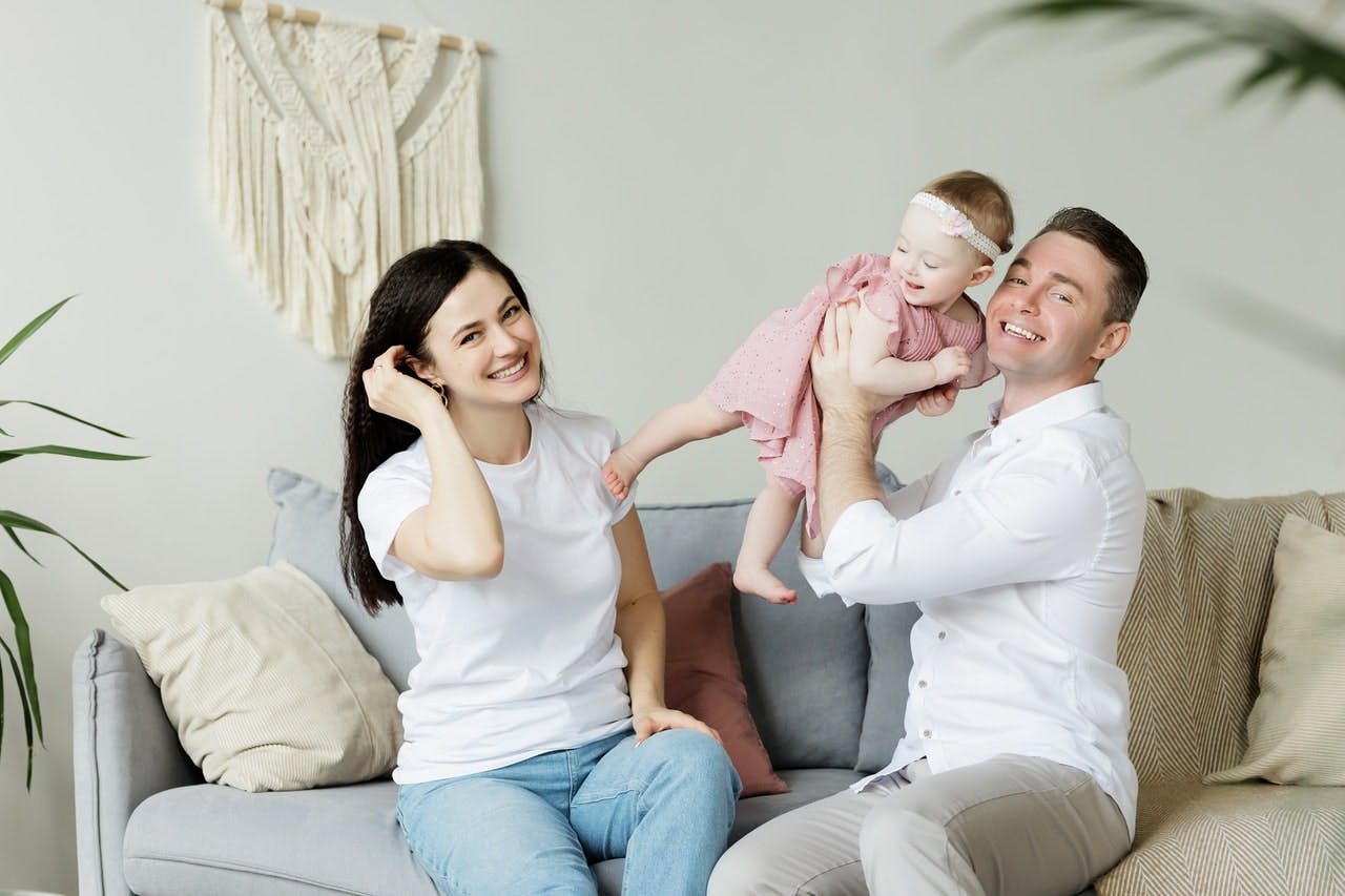 Couple sat on grey sofa play with young child in pink outfit