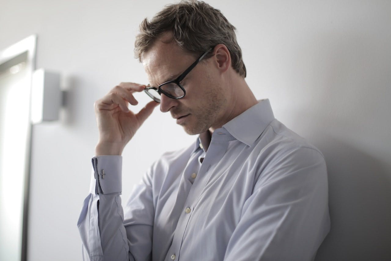 A man wearing glasses and a white shirt, leans against a wall looking worried 