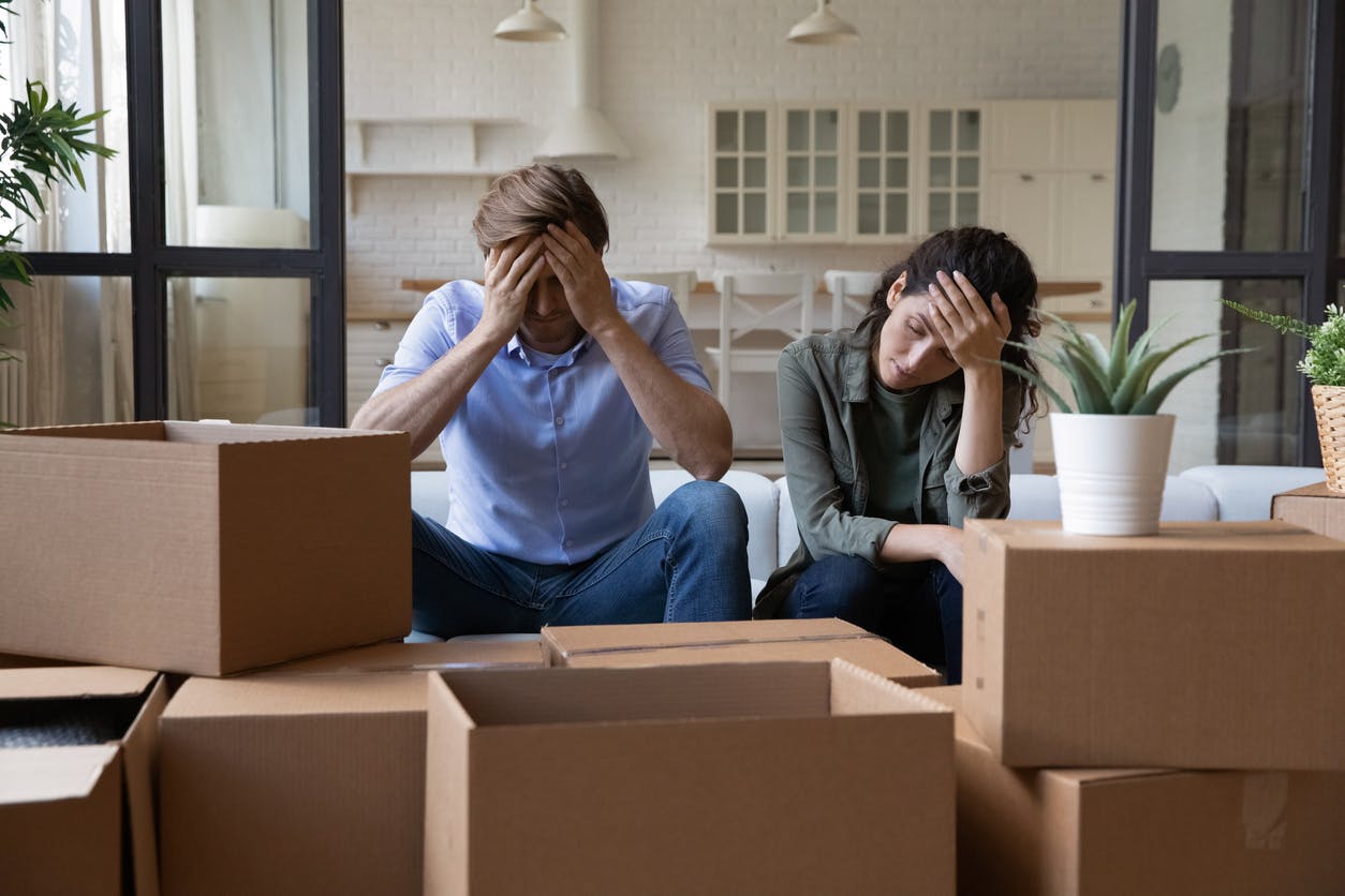A couple holding their heads in a room filled with cardboard boxes.