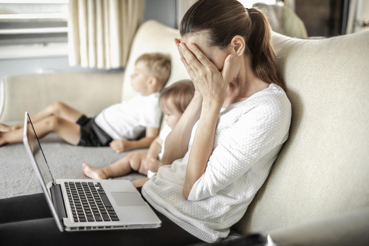Woman sat on a couch with a laptop on her knees and two children in the background.