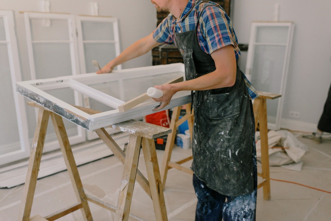 person in apron and check shirt painting window frames resting on wooden trestles in a garage