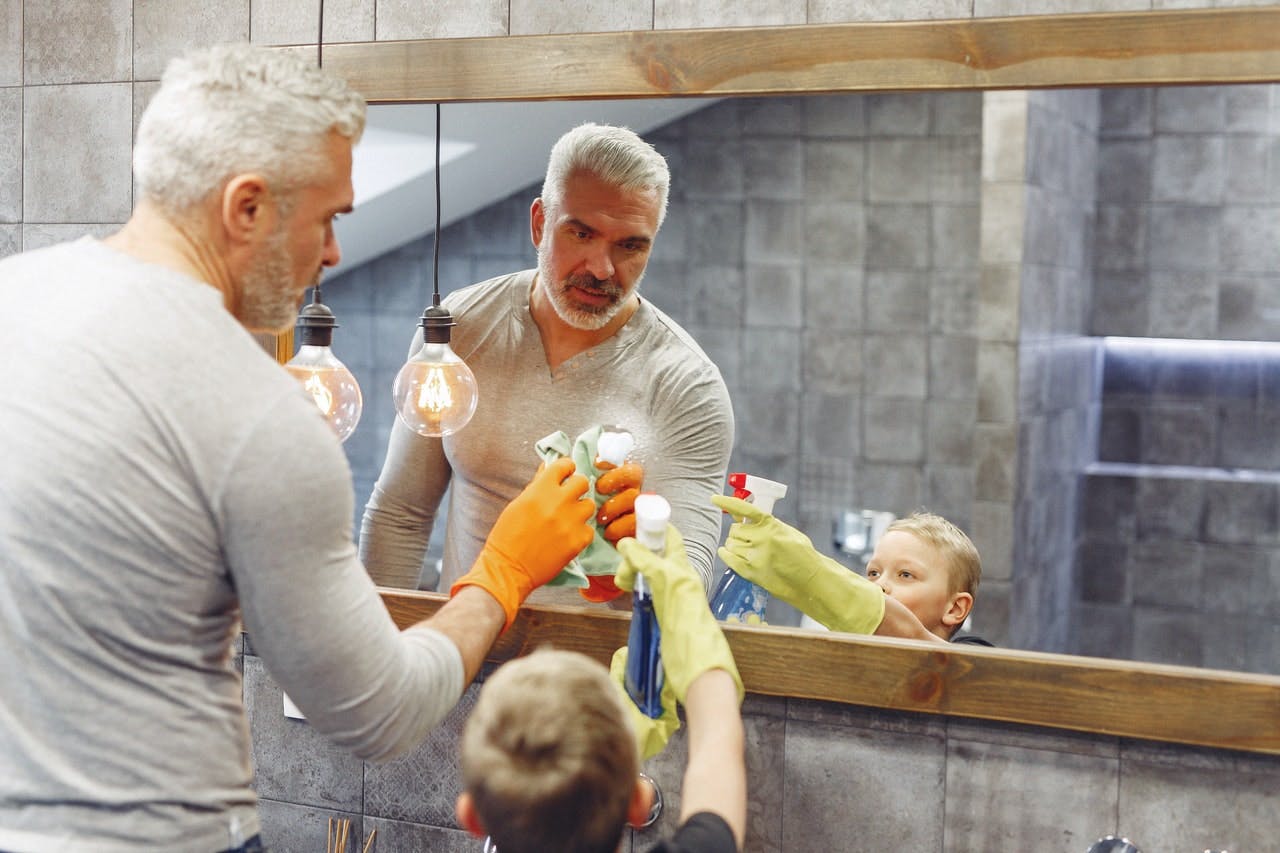 Older man in grey top teaches young child wearing rubber gloves to clean a mirror in a modern bathroom