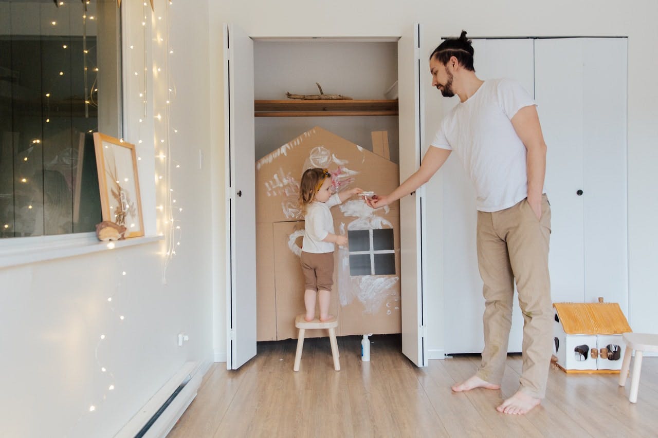 A toddler paints a cardboard cut out of a house, a father helps