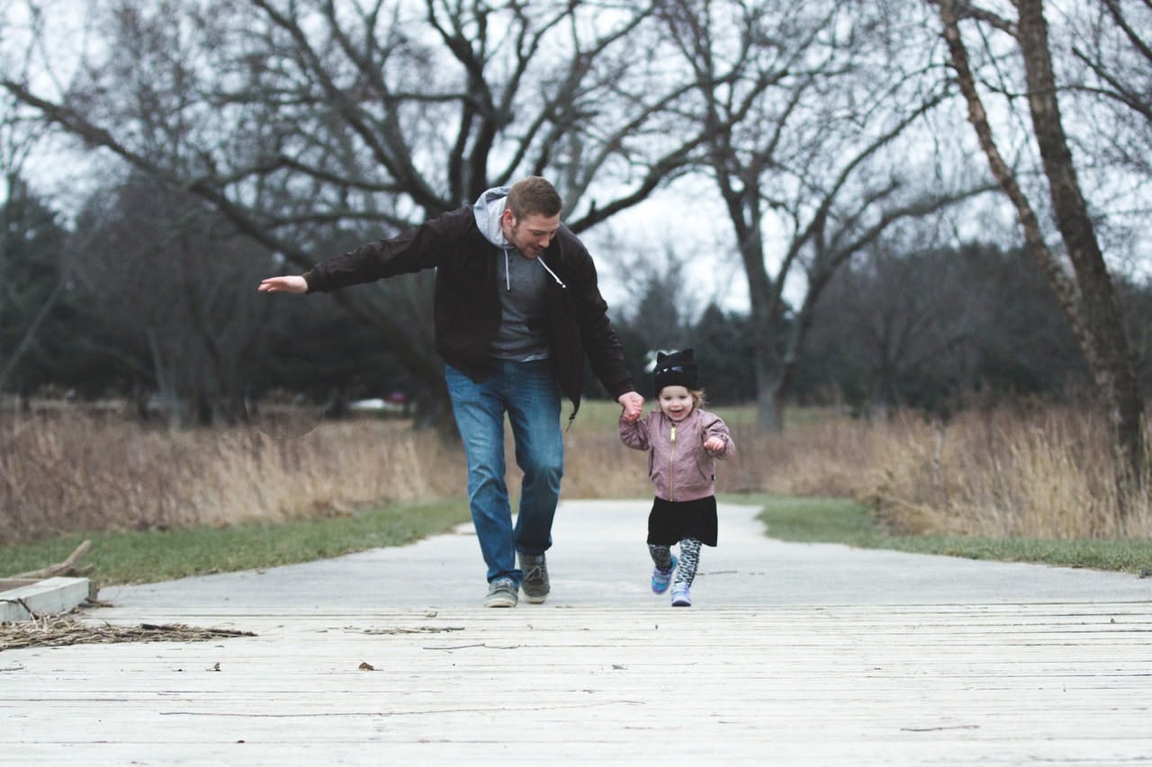 Parent and young child wearing warm clothing run excitedly through a park 