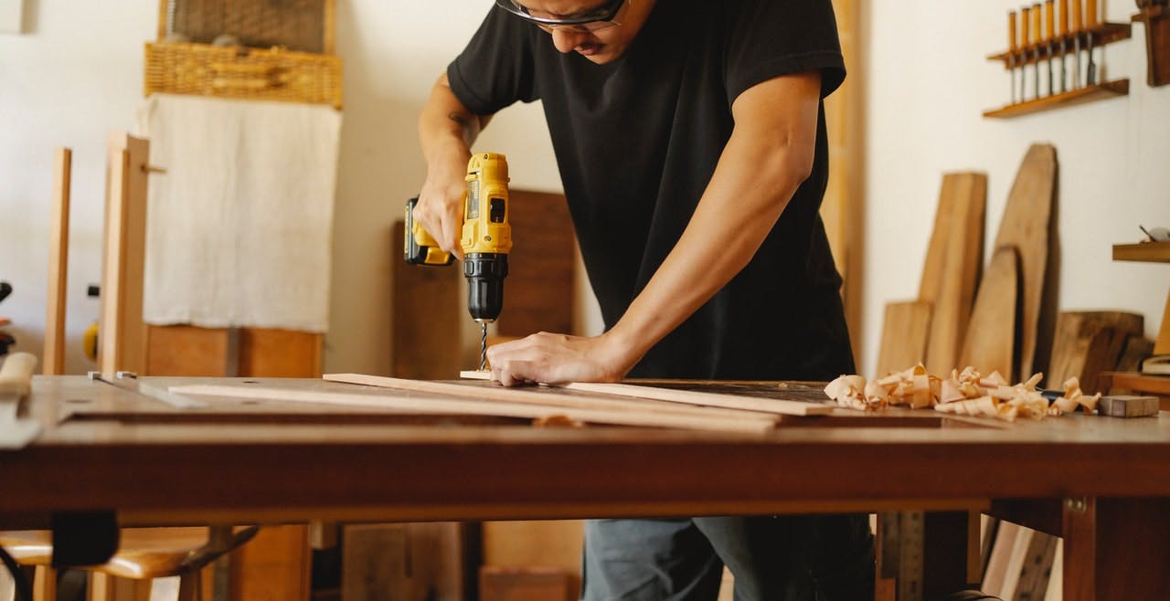 Person wearing protective googles and black t-shirt drills holes in pieces of wood resting on wooden work bench 