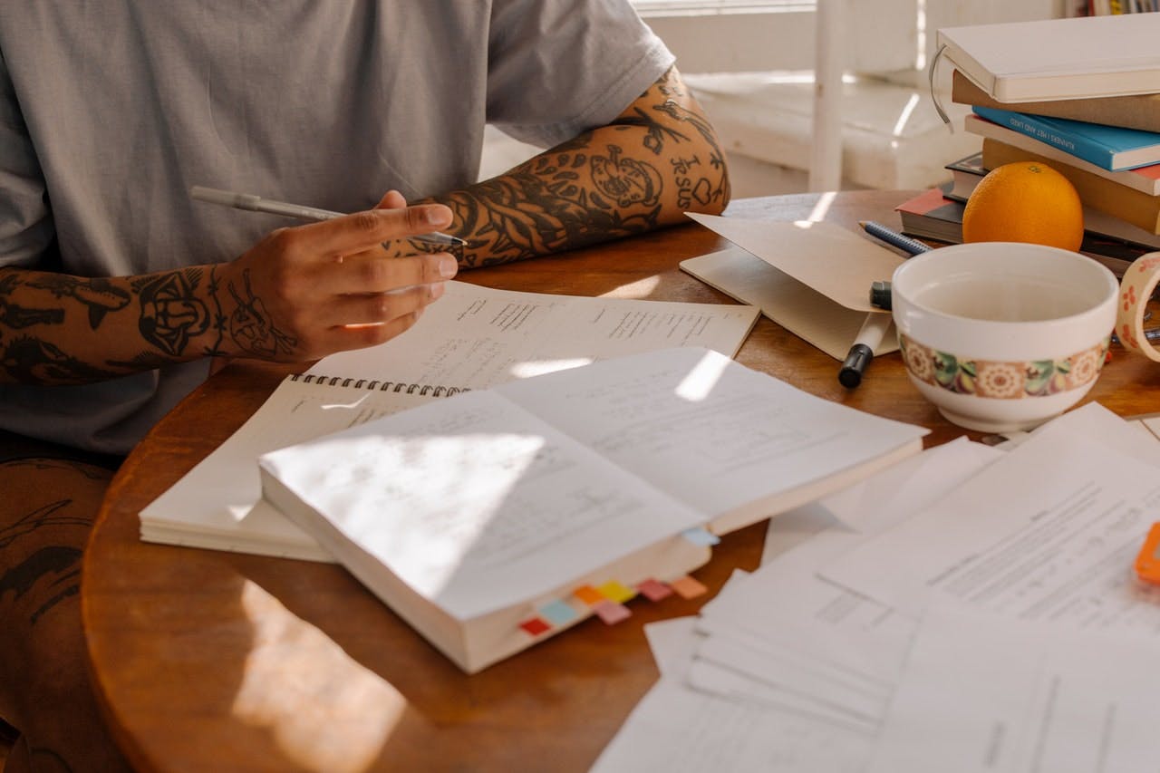 Person with tattoos sits at wooden table, covered in papers and books, studying and making notes 