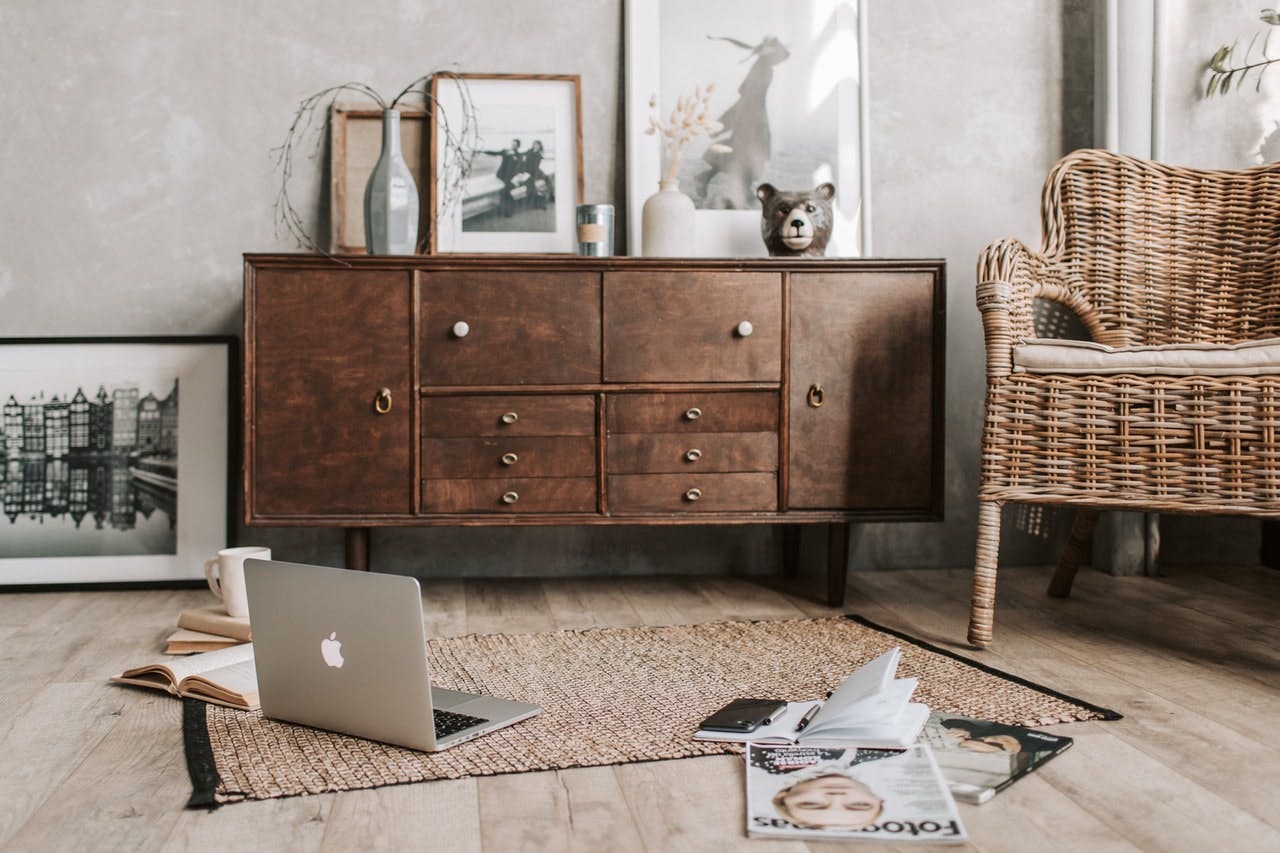 magazines and laptop on woven rug on living room floor, furnished with wooden sideboard and rattan chair, grey walls, and a range of photo print posters 