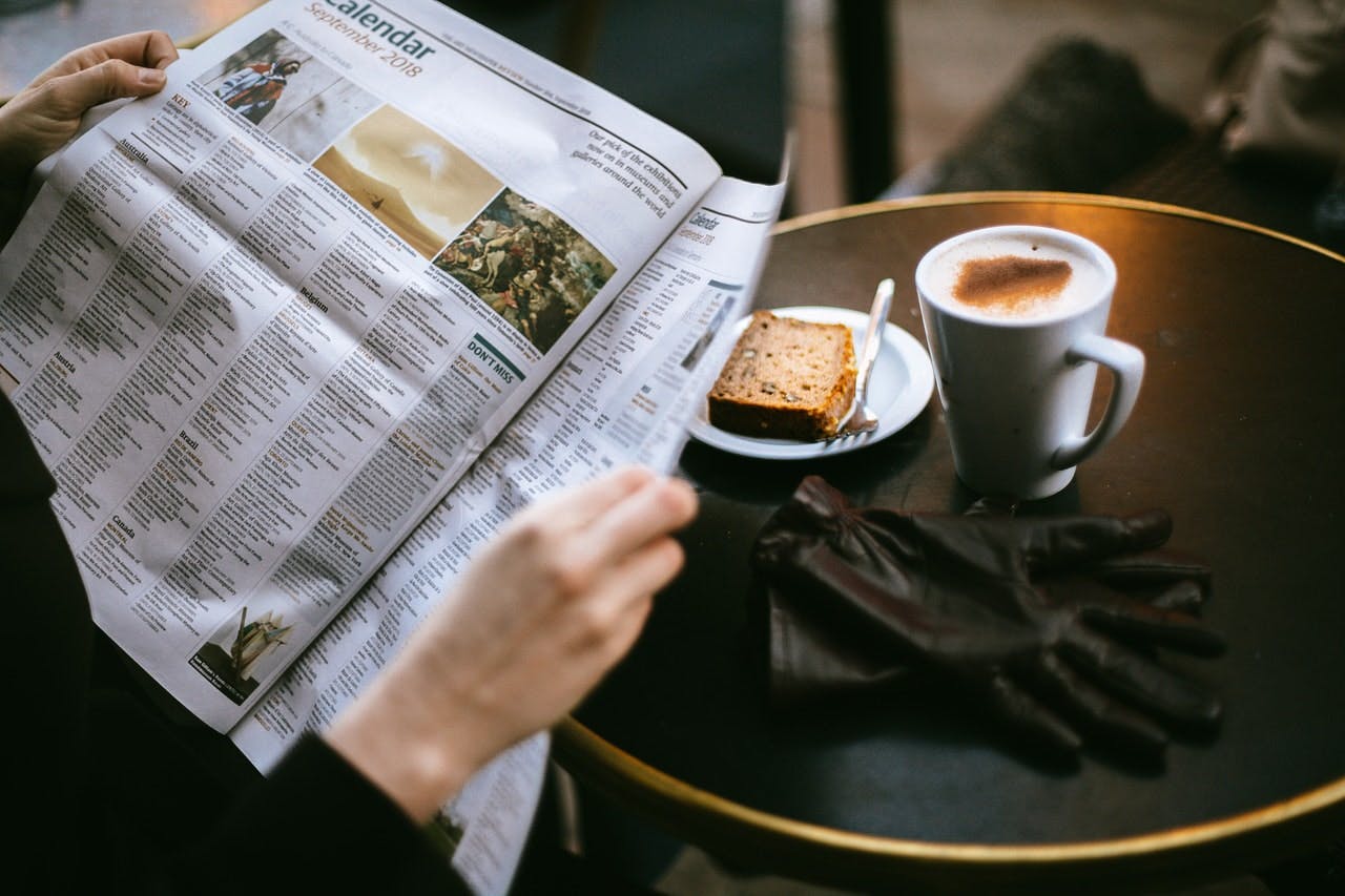 Person reading newspaper at modern cafe table with a cappuccino and slice of cake