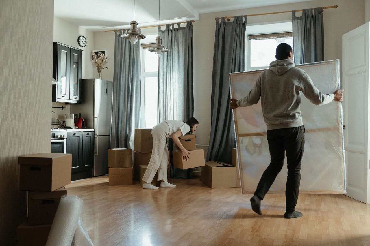 Woman in white t-shirt lifting cardboard moving boxes, while other male home mover in hoodie carries large painting wrapped in bubble wrap