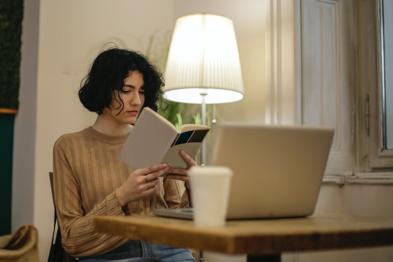 A young tenant reads a book by a table lamp, a laptop and coffee cup on table in front
