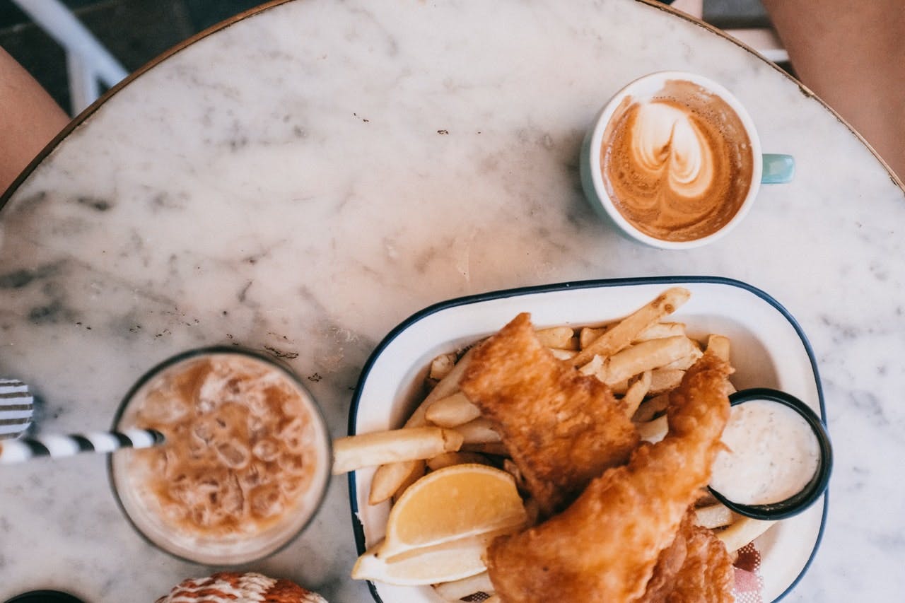 plate of fish and chips, an ice coffee, and a cappuccino on a small marble table 