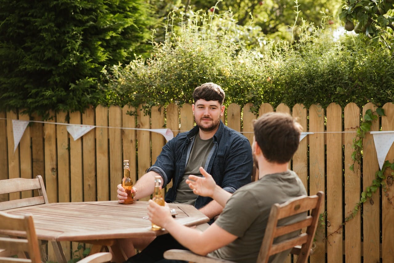 Two neighbours drink a beer in a sunny garden, a wooden fence behind has bunting on it