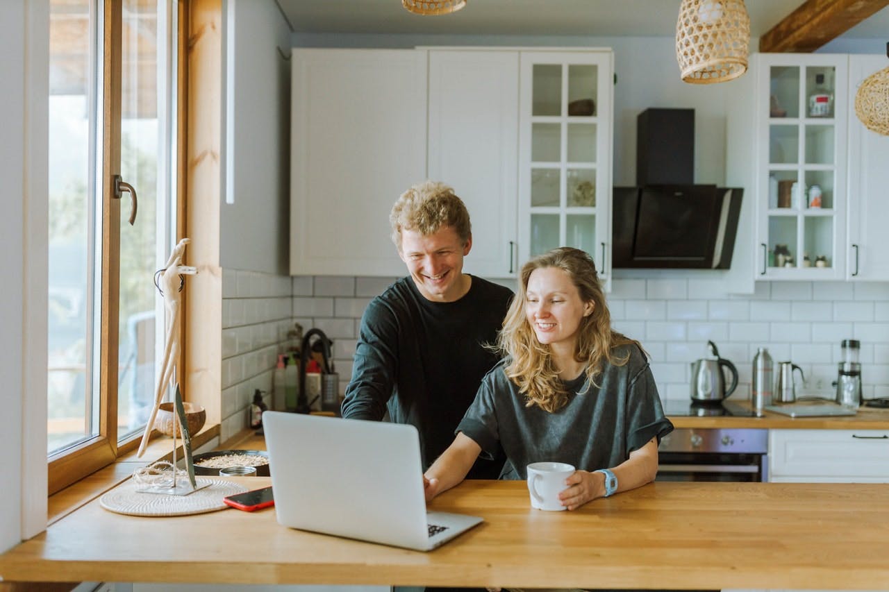 A smiling couple sat in a kitchen with modern white and wood fixtures look at laptop